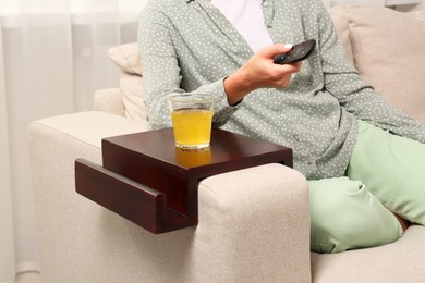 Glass of juice on wooden sofa armrest table. Woman holding remote control at home, closeup