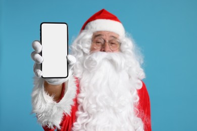 Photo of Merry Christmas. Santa Claus showing smartphone on light blue background, selective focus. Mockup for design