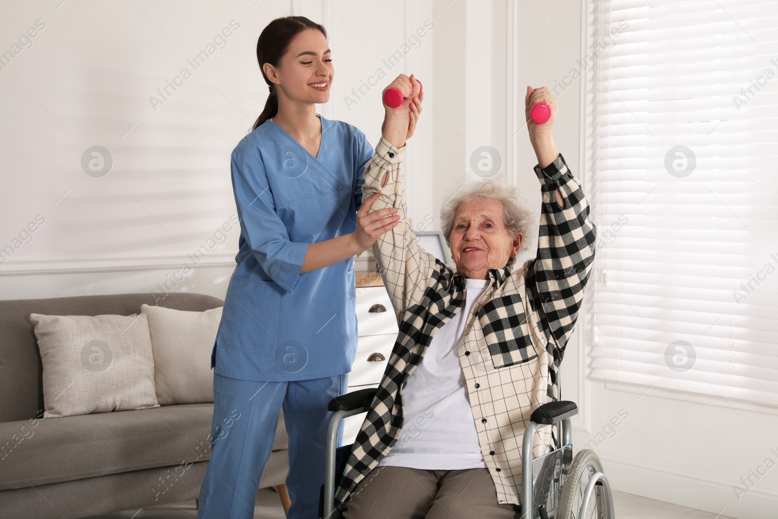 Photo of Senior woman in wheelchair doing physical exercise and young caregiver helping her indoors. Home health care service