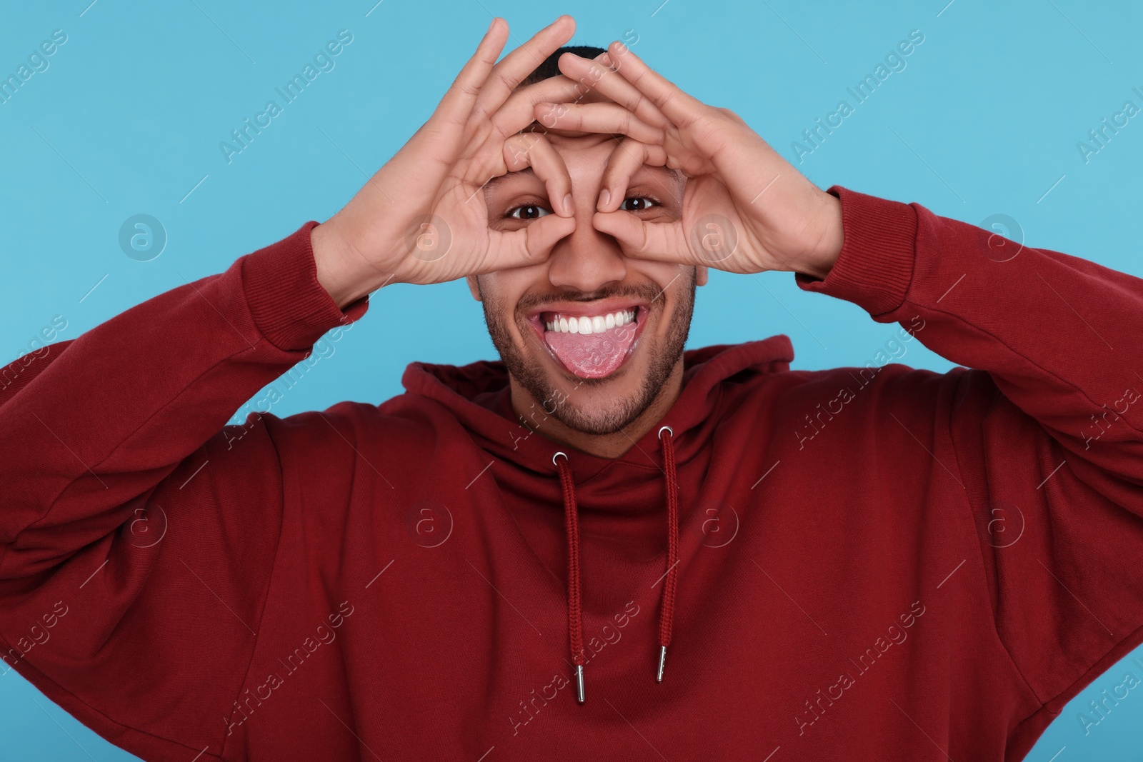 Photo of Happy young man showing his tongue and hand glasses gesture on light blue background