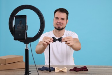 Photo of Smiling fashion blogger showing bow ties while recording video at table against light blue background