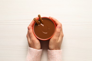 Woman holding cup of yummy hot chocolate with cinnamon at white wooden table, top view