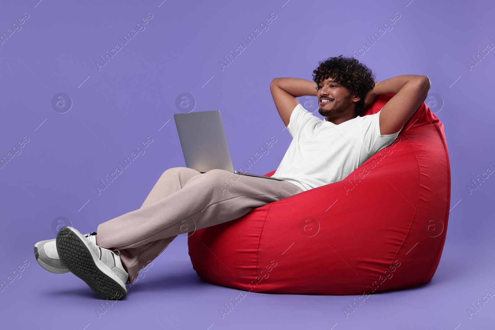 Photo of Smiling man with laptop sitting on beanbag chair against purple background