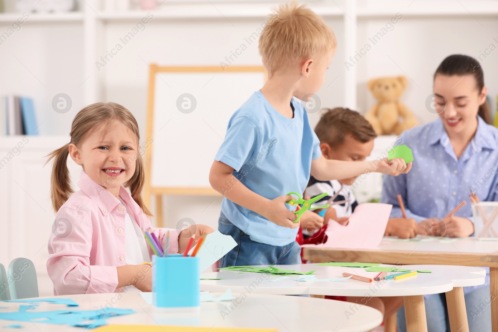 Photo of Nursery teacher and group of cute little children making toys from color paper at desks in kindergarten. Playtime activities