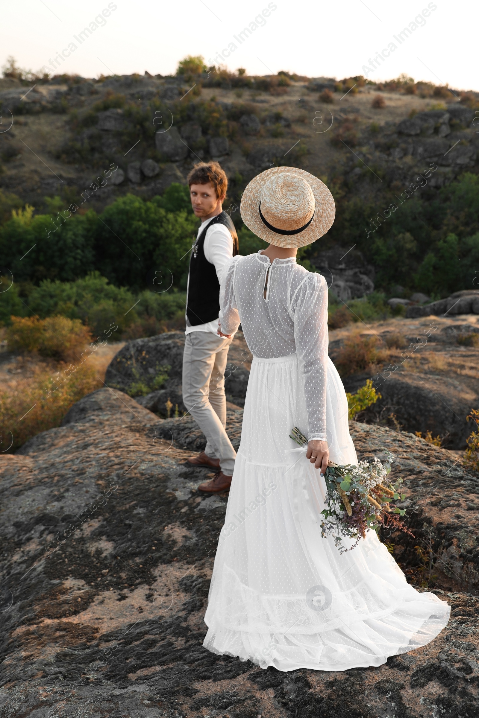 Photo of Happy newlyweds with beautiful field bouquet standing on rock outdoors