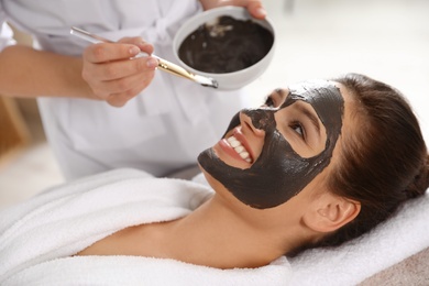 Cosmetologist applying black mask onto woman's face in spa salon