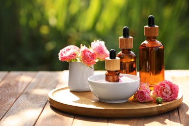 Bottles of rose essential oil and flowers on wooden table outdoors, space for text