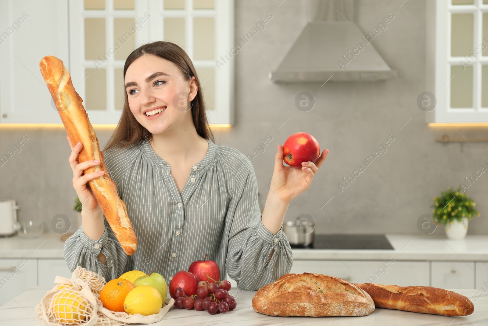 Photo of Woman with baguette, apple and string bag of fresh fruits at light marble table in kitchen