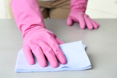 Photo of Woman in gloves wiping grey table with rag indoors, closeup