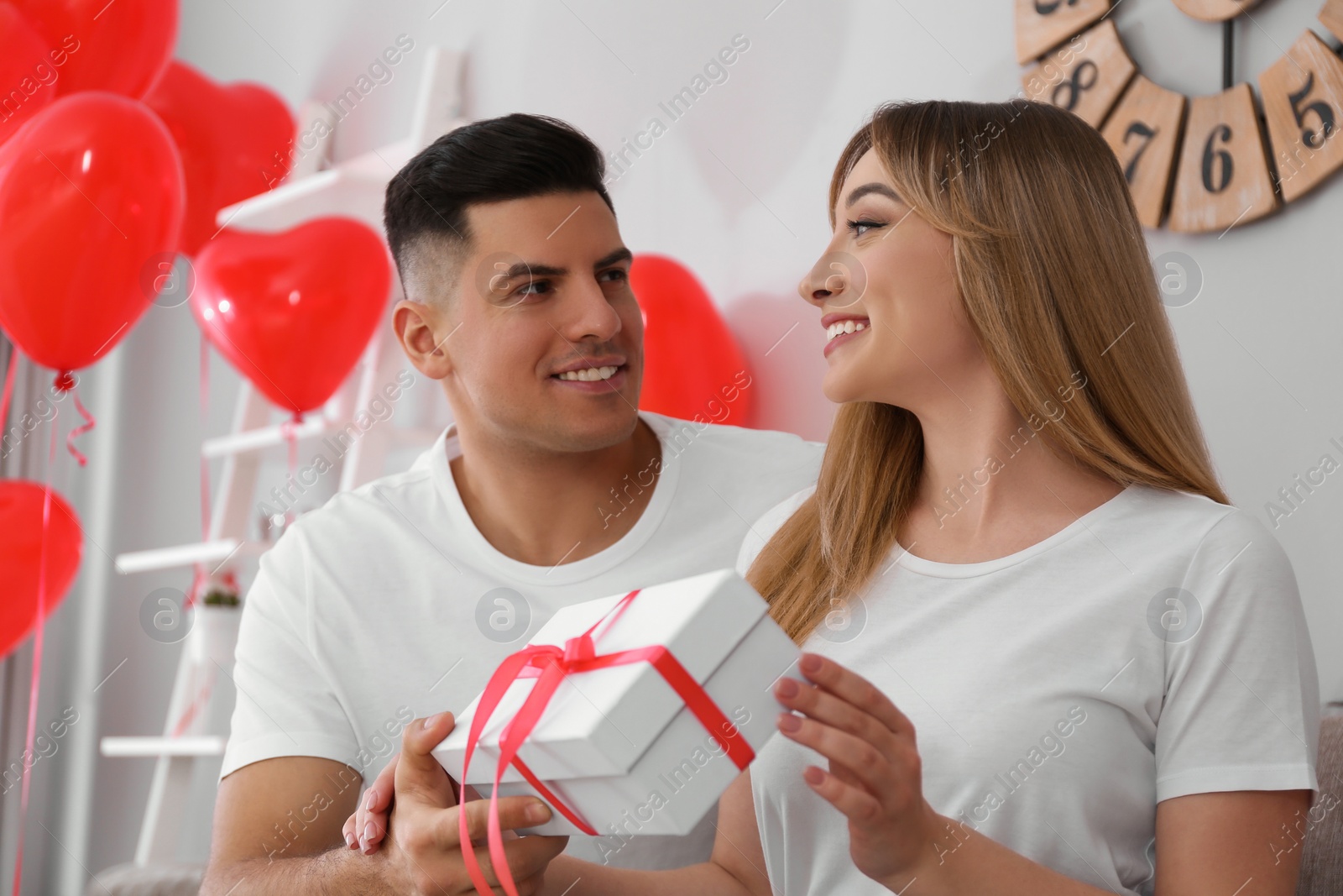 Photo of Man presenting gift to his girlfriend in room decorated with heart shaped balloons. Valentine's day celebration