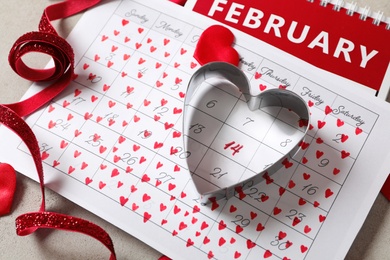 Photo of Calendar with marked Valentine's Day, red ribbon and heart shaped cookie cutter on grey table, closeup