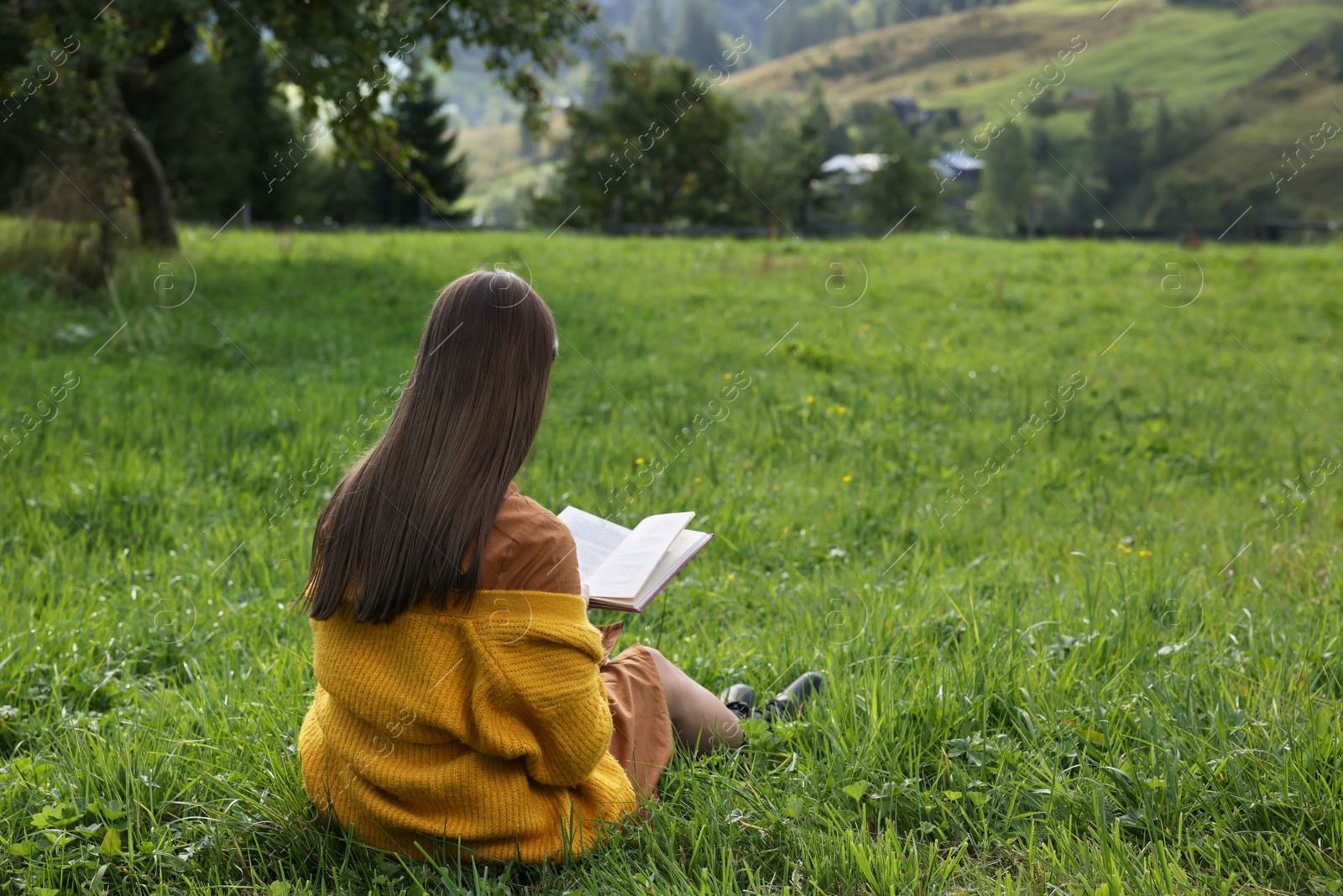 Photo of Young woman reading book on green meadow, back view