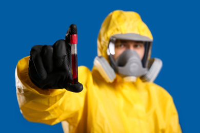 Photo of Man in chemical protective suit holding test tube of blood sample against blue background, focus on hand. Virus research