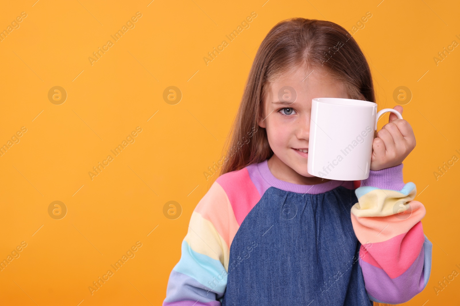 Photo of Happy girl covering eye with white ceramic mug on orange background, space for text