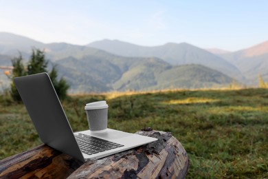 Photo of Modern laptop and coffee cup on log in mountains, space for text. Working outdoors