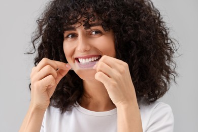 Young woman applying whitening strip on her teeth against light grey background