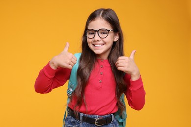 Photo of Cute schoolgirl in glasses showing thumbs up on orange background