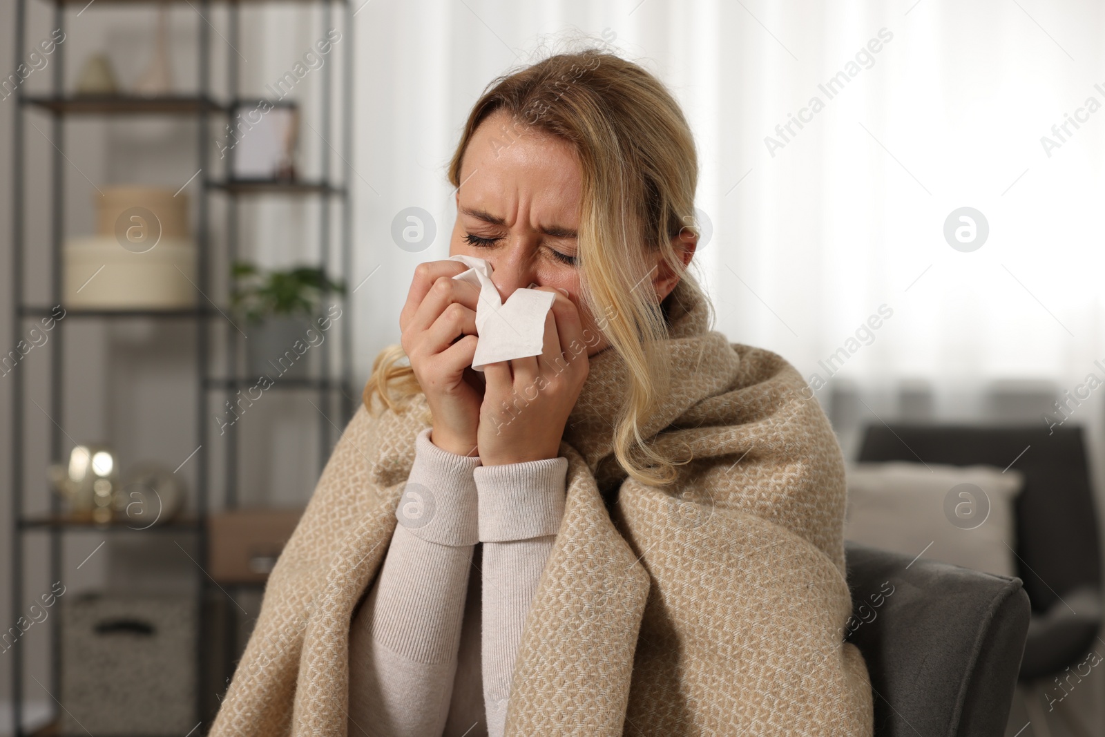 Photo of Sick woman wrapped in blanket blowing nose in tissue at home. Cold symptoms