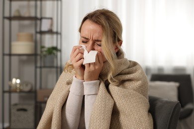 Photo of Sick woman wrapped in blanket blowing nose in tissue at home. Cold symptoms