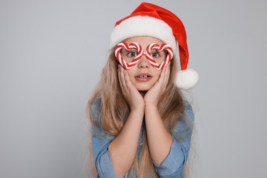 Girl wearing decorative Christmas eyeglasses in shape of hearts and Santa hat on light grey background