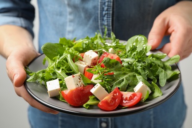 Photo of Woman holding plate of delicious salad with feta cheese, arugula and tomatoes on light background, closeup