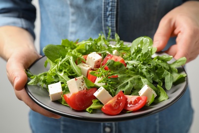 Woman holding plate of delicious salad with feta cheese, arugula and tomatoes on light background, closeup