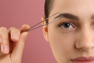 Photo of Eyebrow correction. Young woman with tweezers on pink background, closeup