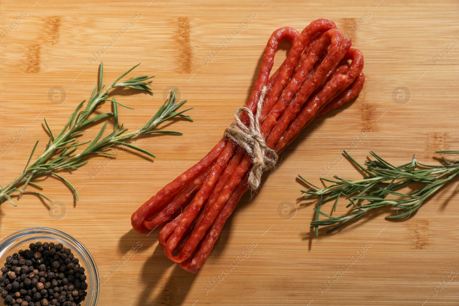 Photo of Bundle of delicious kabanosy with peppercorn and rosemary on wooden table, flat lay