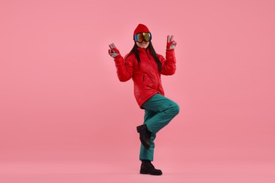 Photo of Happy woman in winter sportswear and goggles showing peace sign on pink background