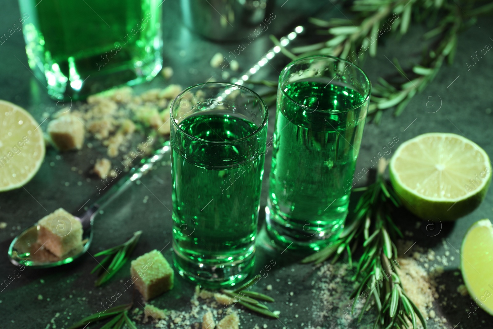 Photo of Absinthe in shot glasses, spoon, brown sugar, lime and rosemary on gray table, closeup. Alcoholic drink