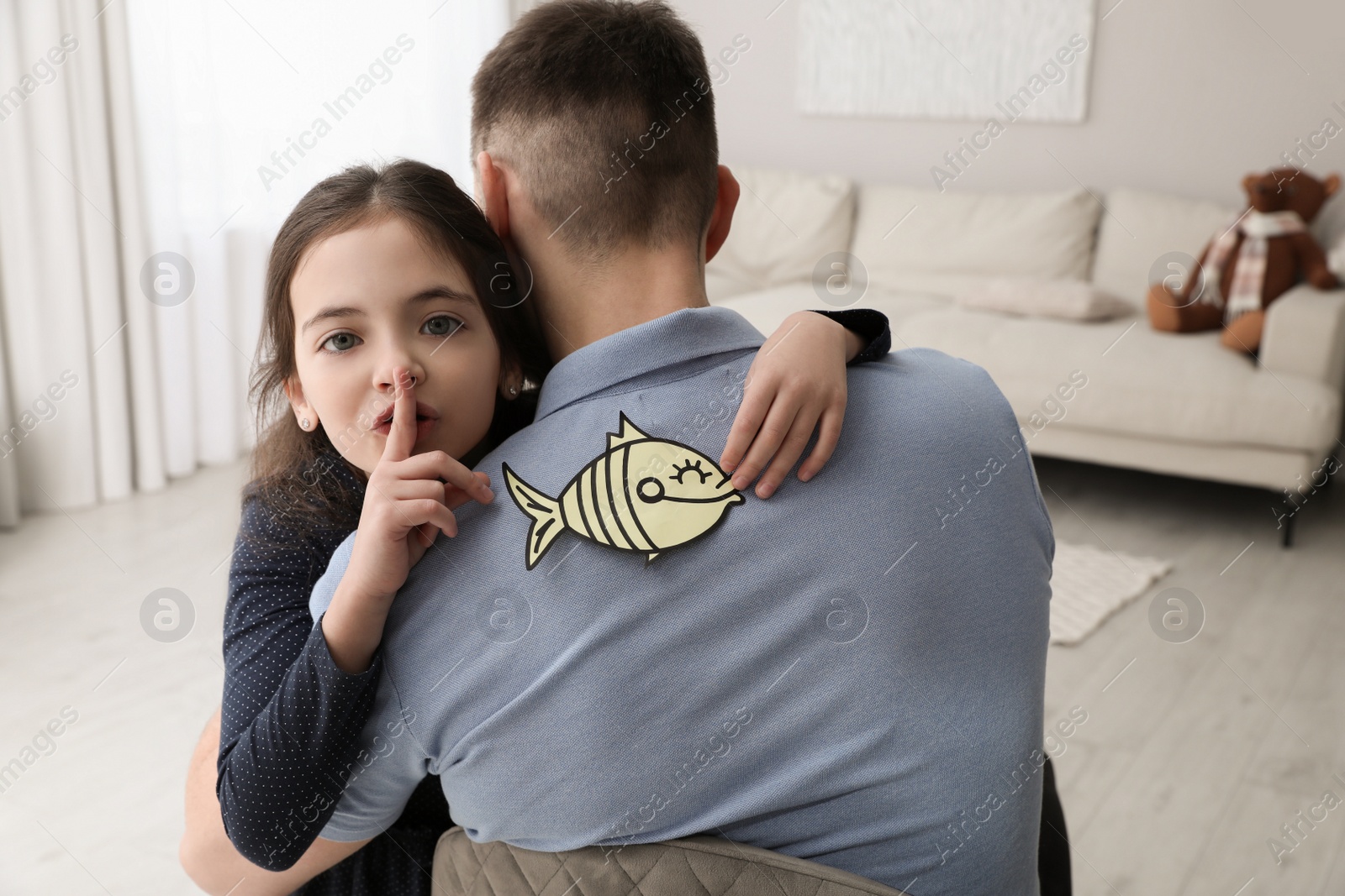 Photo of Cute little girl sticking paper fish to father's back at home