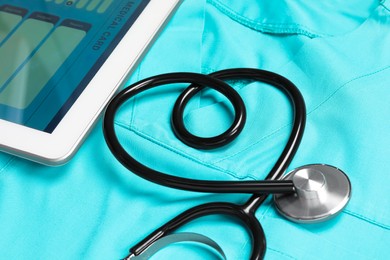 Photo of Stethoscope in shape of heart and tablet on turquoise medical uniform, closeup