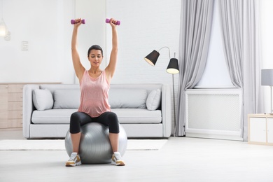 Photo of Young woman doing exercise with dumbbells on fitness ball at home. Space for text