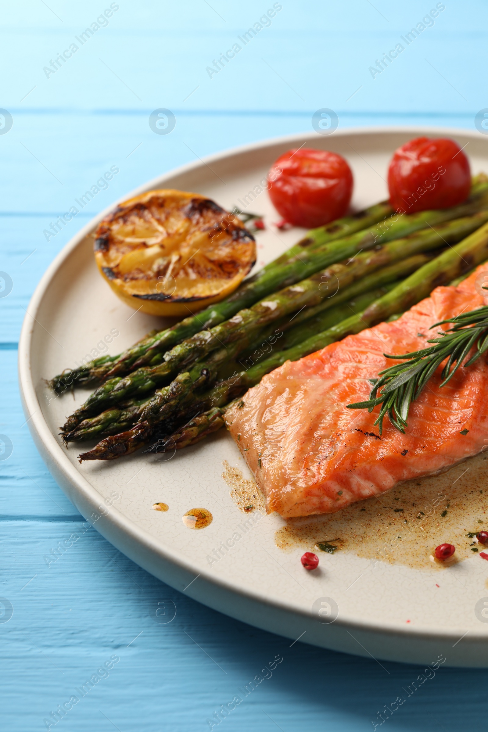 Photo of Tasty grilled salmon with tomatoes, asparagus and spices on light blue wooden table, closeup