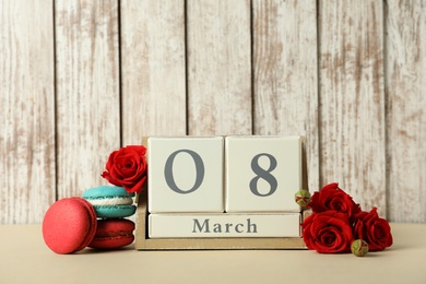 Wooden block calendar with date 8th of March, macarons and roses on table against light background, space for text. International Women's Day