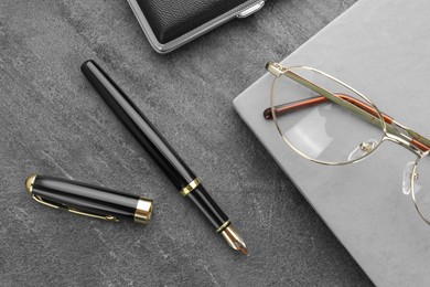 Photo of Stylish black fountain pen, glasses, notebook and cigarette case on grey textured table, flat lay