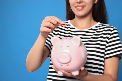 Photo of Young woman putting coin into piggy bank on light blue background, closeup