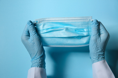 Photo of Doctor in latex gloves holding disposable face mask on light blue background, closeup. Protective measures during coronavirus quarantine