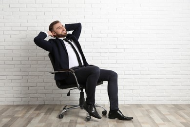 Young man sitting in office chair near white brick wall indoors, space for text