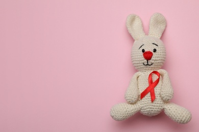 Photo of Cute knitted toy bunny with red ribbon on pink background, top view and space for text. AIDS disease awareness