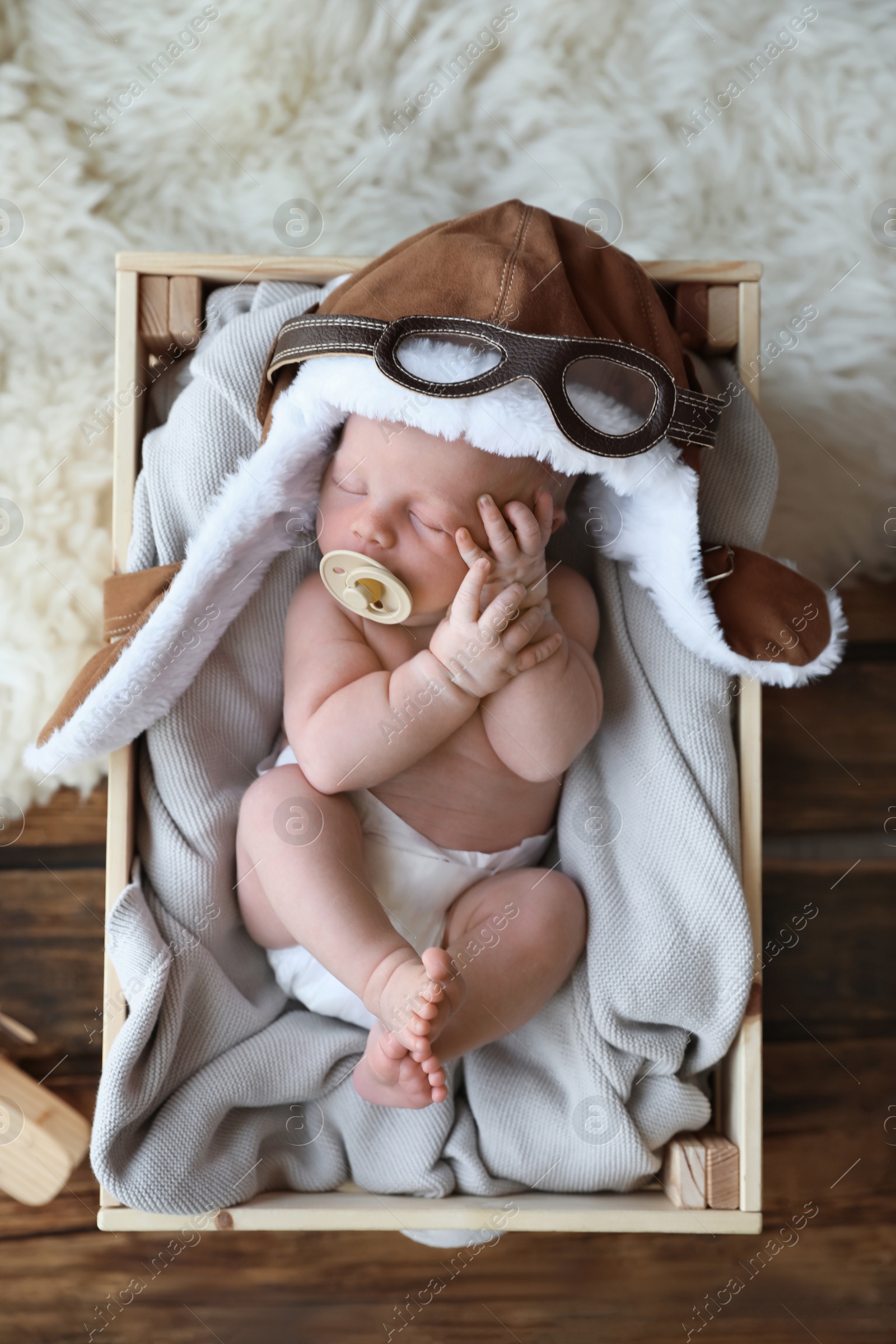 Photo of Cute newborn baby wearing aviator hat with toy sleeping in wooden crate, top view