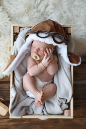 Photo of Cute newborn baby wearing aviator hat with toy sleeping in wooden crate, top view
