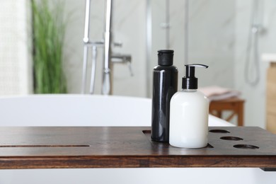 Photo of Wooden bath tray with bottles of shower gels on tub indoors, space for text