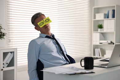 Man with fake eyes painted on sticky notes snoozing at workplace in office