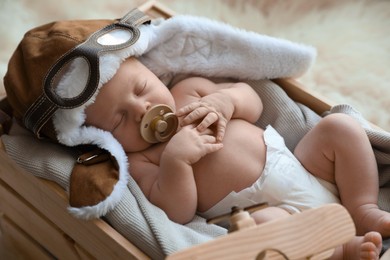 Cute newborn baby wearing aviator hat with toy sleeping in wooden crate