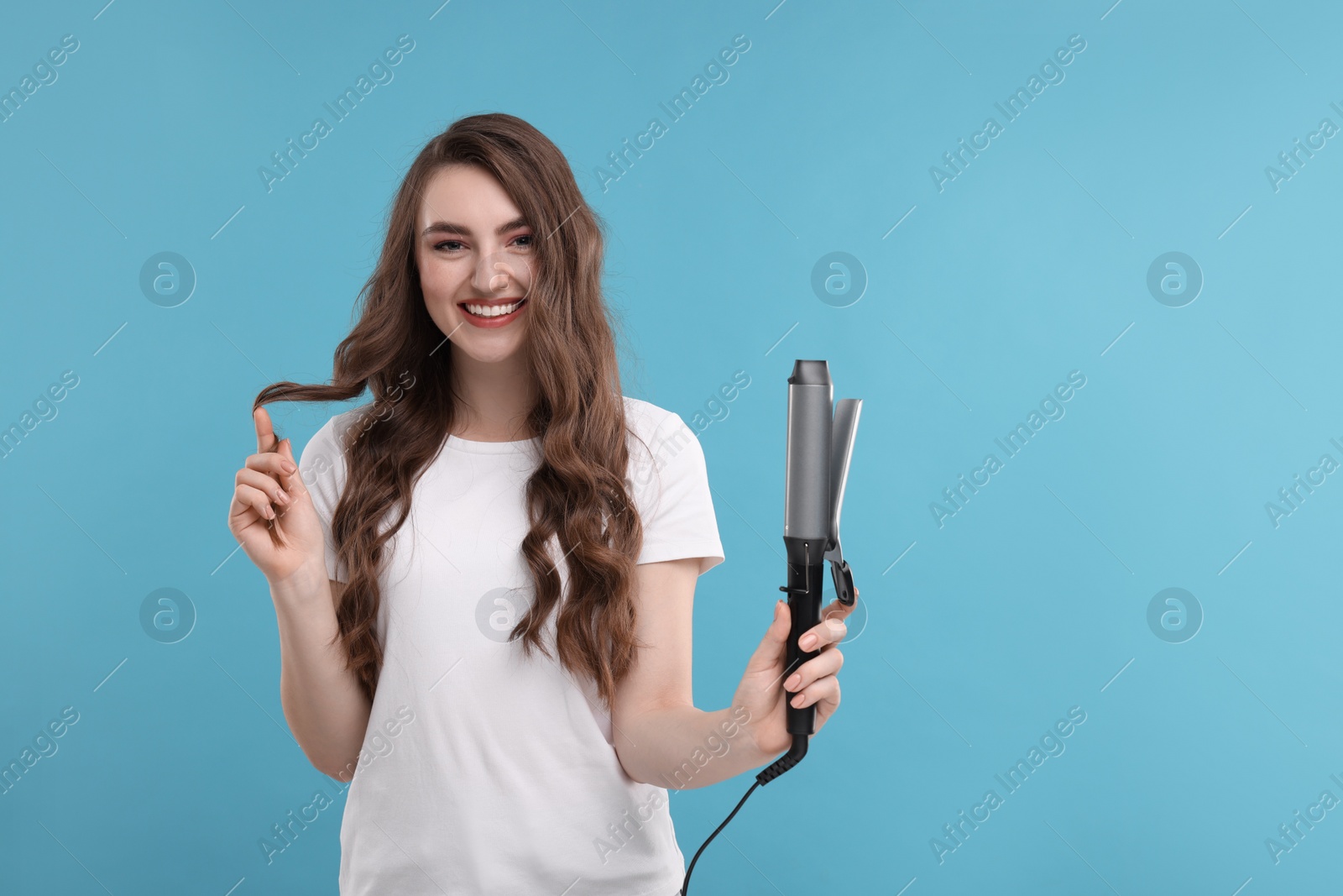 Photo of Happy young woman with beautiful hair holding curling iron on light blue background, space for text