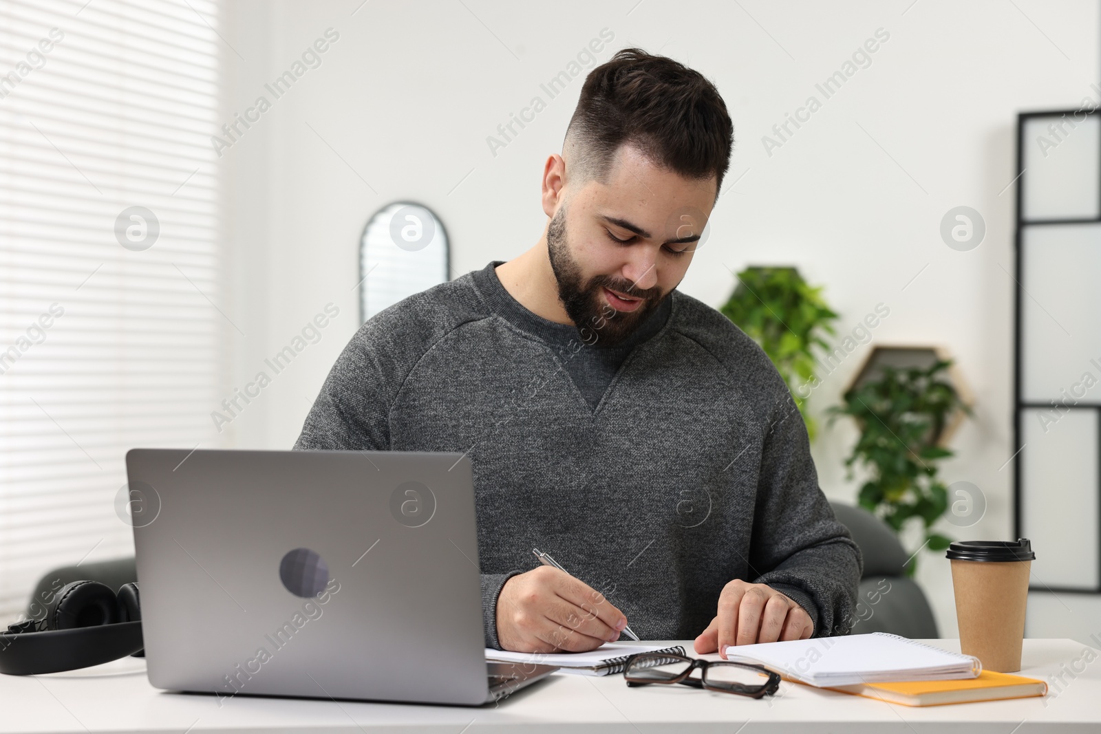 Photo of E-learning. Young man taking notes during online lesson at white table indoors