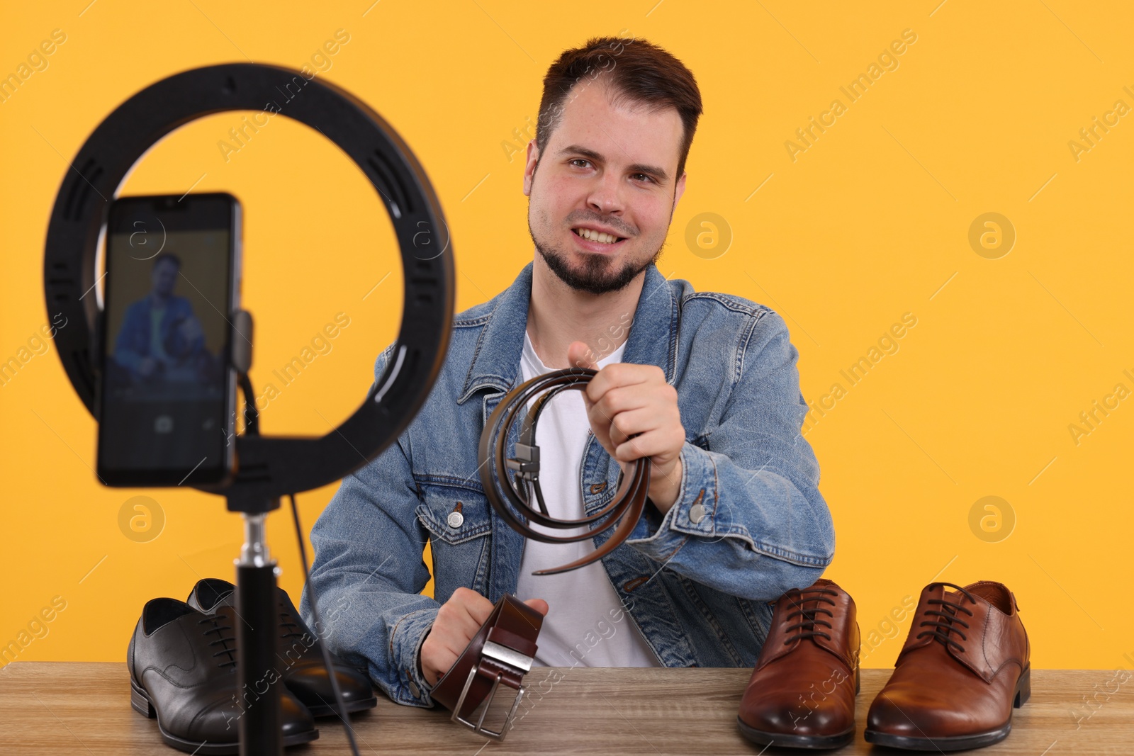 Photo of Smiling fashion blogger showing men's accessories while recording video at table against orange background