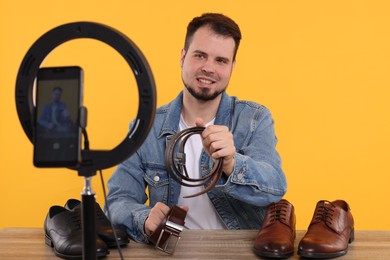 Smiling fashion blogger showing men's accessories while recording video at table against orange background