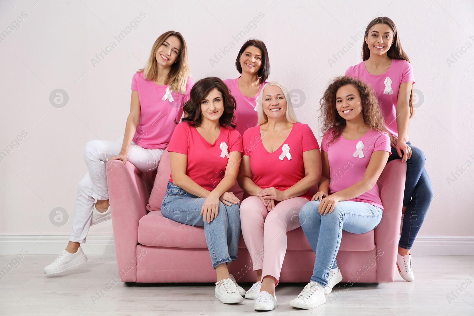 Photo of Group of women with silk ribbons sitting on sofa against light wall. Breast cancer awareness concept